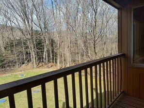 A wooden deck with a railing overlooking a grassy area and a forested landscape. - Cozy Mountain Getaway - Ski, Hike, Golf & Unwind (Waitsfield)
