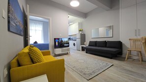 A living room with a sofa, a television, a washing machine, and a dining area. - KYANA Luxury KVA (Kavala)