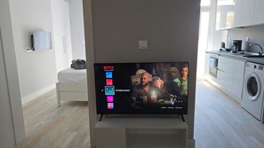 Ein modernes Wohnzimmer mit Flachbildfernseher, der einen Film zeigt, einem ordentlich bezogenen Bett und einer Küchenzeile mit Geräten.