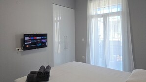 A hotel room with a bed, a wall-mounted TV displaying streaming services, a window with sheer curtains, and a ceiling light. - KYANA Luxury KVA (Kavala)