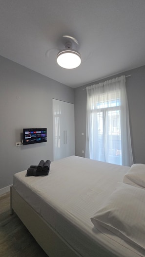 A hotel room with a bed, a wall-mounted TV displaying streaming services, a window with sheer curtains, and a ceiling light. - KYANA Luxury KVA (Kavala)