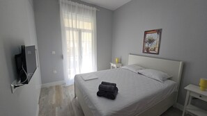 Habitación de hotel con cama, televisión, ventana con cortinas, mesita de noche con lámpara y un cuadro en la pared.