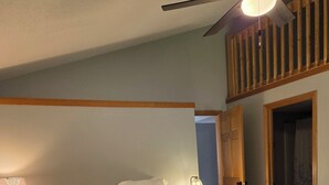 A bedroom with a bed, a red throw blanket, a nightstand with a lamp, and a wooden ceiling fan.