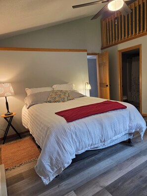 Ein Schlafzimmer mit einem Bett, einer roten Tagesdecke, einem Nachttisch mit Lampe und einem hölzernen Deckenventilator.
