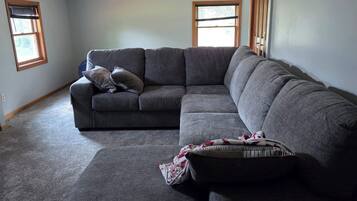 A living room with a sectional sofa, a small ottoman, and a ceiling fan. There are two windows with blinds.