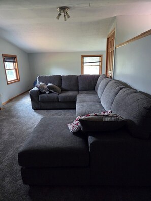 A living room with a sectional sofa, a small ottoman, and a ceiling fan. There are two windows with blinds.
