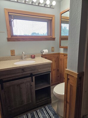 A bathroom with a wooden vanity, a toilet, a window with blinds, and a mirror.