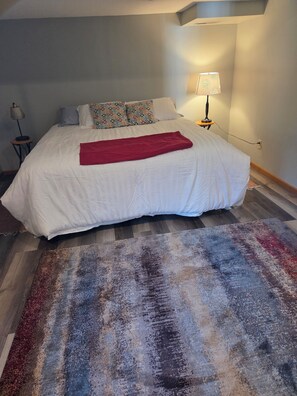A bed with a white comforter, a red throw blanket, and patterned pillows. There is a lamp on a side table next to the bed.