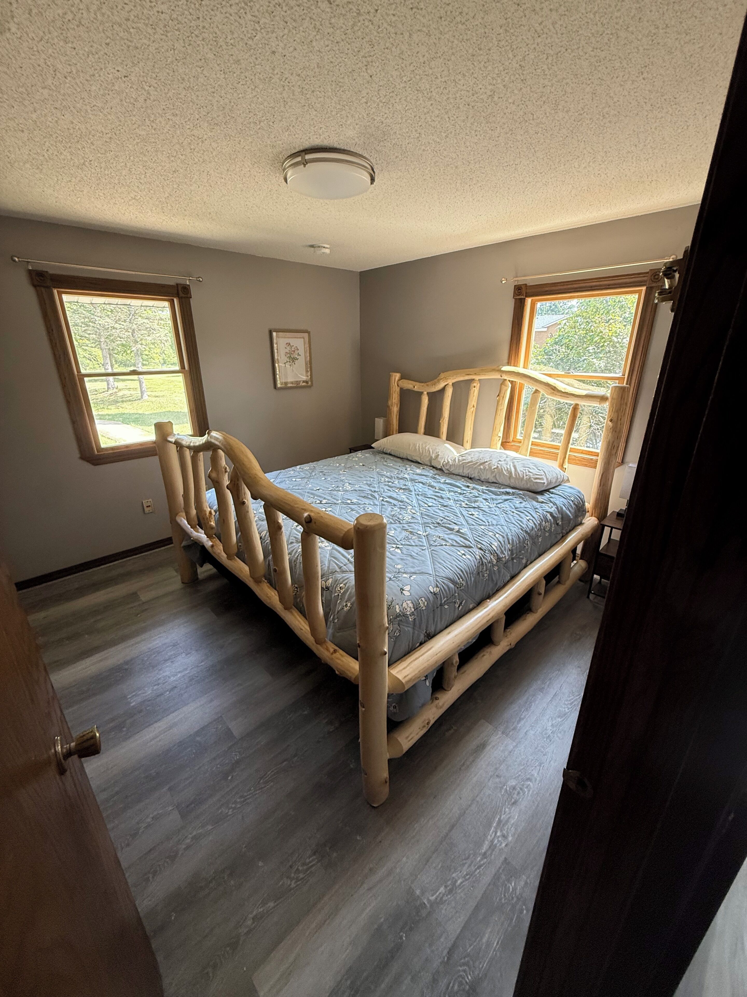 A bedroom with a wooden bed frame, two windows, and a ceiling light.