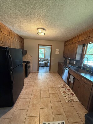 A kitchen with wooden cabinets, a black refrigerator, a tiled floor, and a doorway leading to a living area. - Rush Lake Retreat -plenty of room to sleep, even more room to unwind! (Rush City)