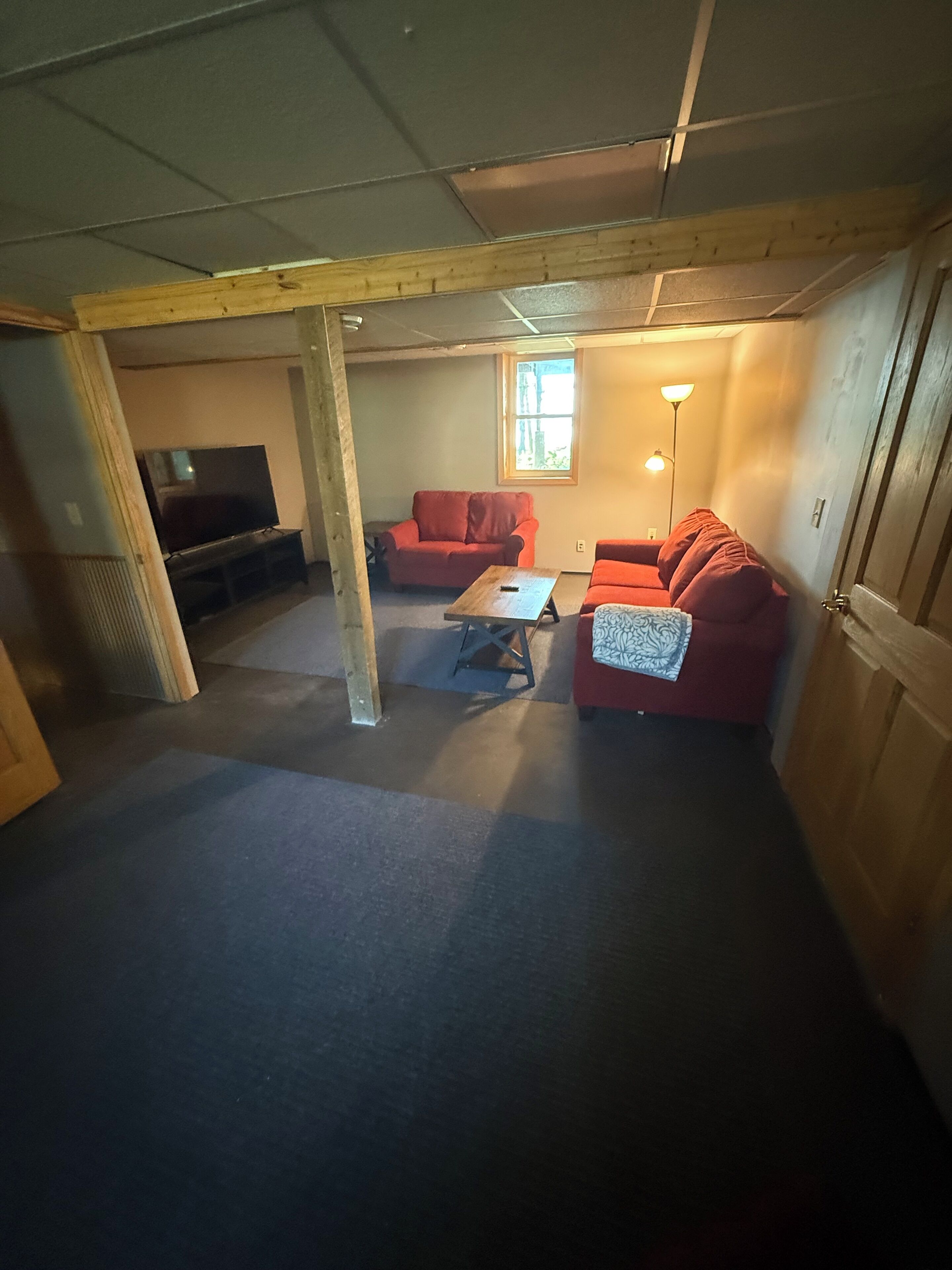 A room with a wooden ceiling, a television, two red sofas, a small table, and a window with natural light.
