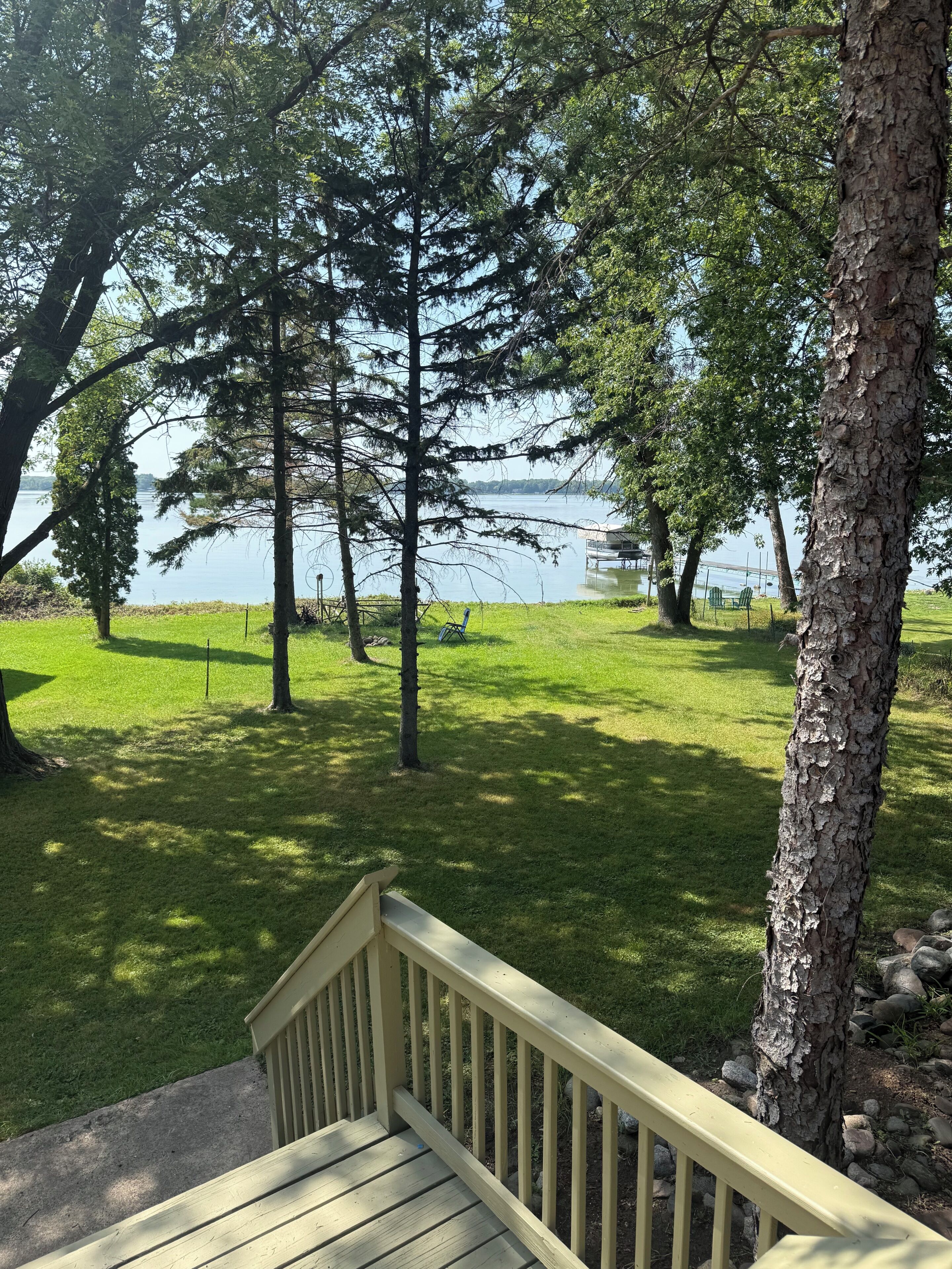 A wooden deck with a railing overlooking a grassy area with trees and a body of water in the background.