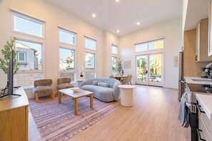 Ein geräumiges Wohnzimmer mit grauem Sofa, Holztisch und einem kleinen Beistelltisch. Ein großes Fenster sorgt für natürliches Licht und bietet einen Ausblick nach draußen.
