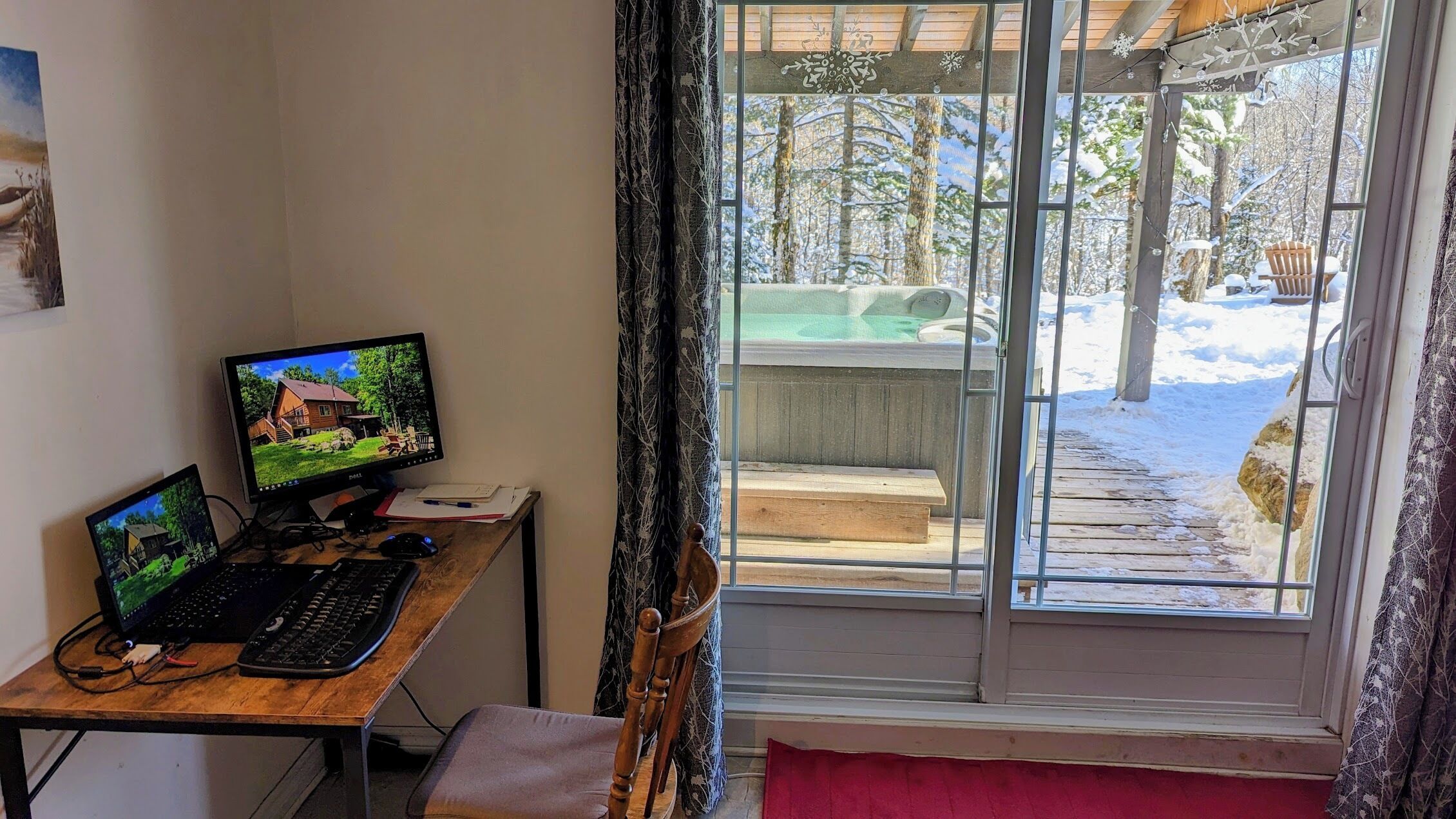 Una habitación con escritorio, dos monitores, teclado y silla. Una ventana con cortinas da a un espacio exterior nevado con un jacuzzi.