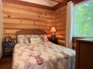 A wooden cabin-style bedroom with a bed, a nightstand, a lamp, and a window with a view of trees. - Le Boisé Chalet: Spa and River (Brownsburg)