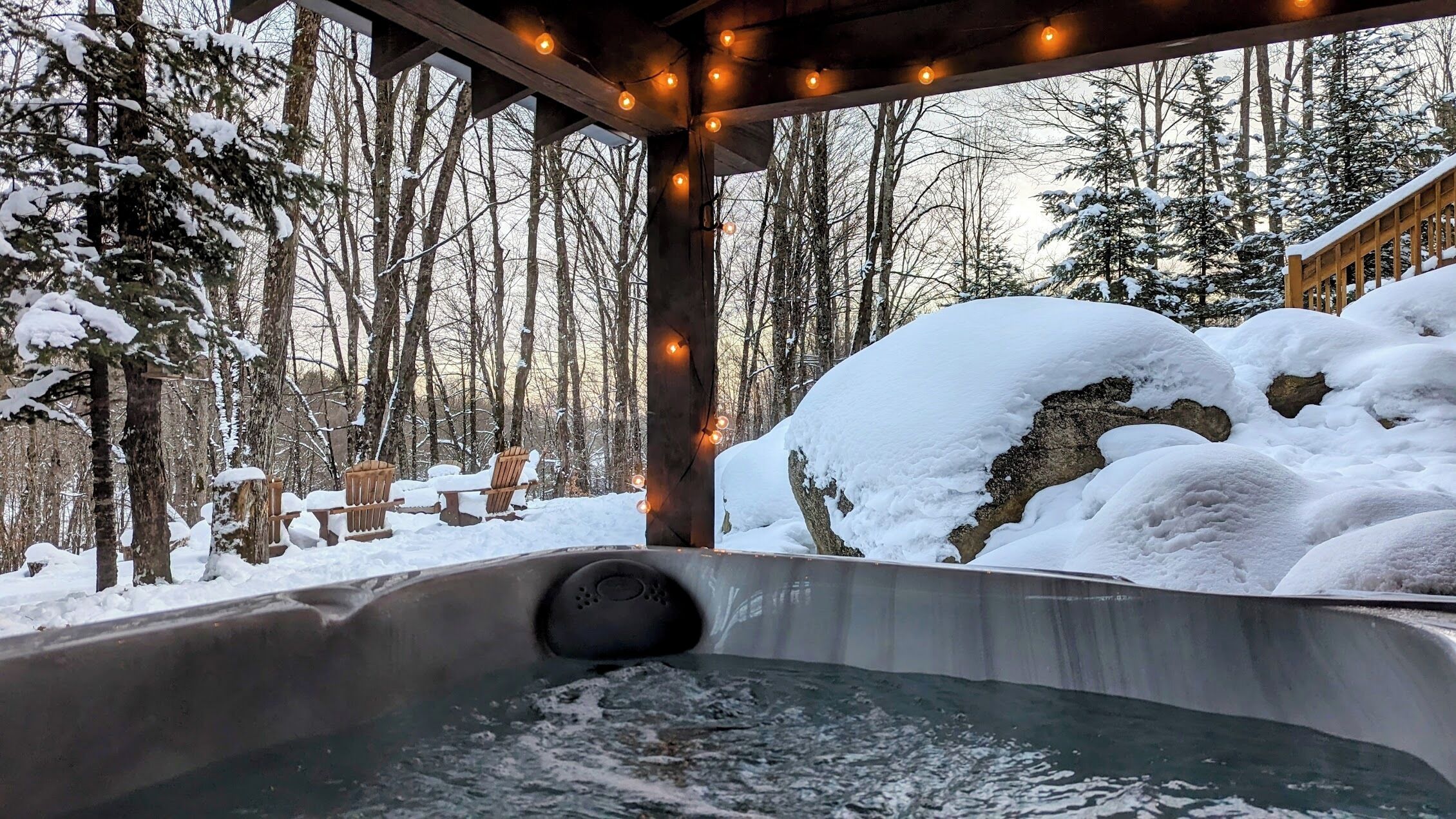 A hot tub with a wooden deck and string lights, surrounded by snow-covered trees and a stone wall.