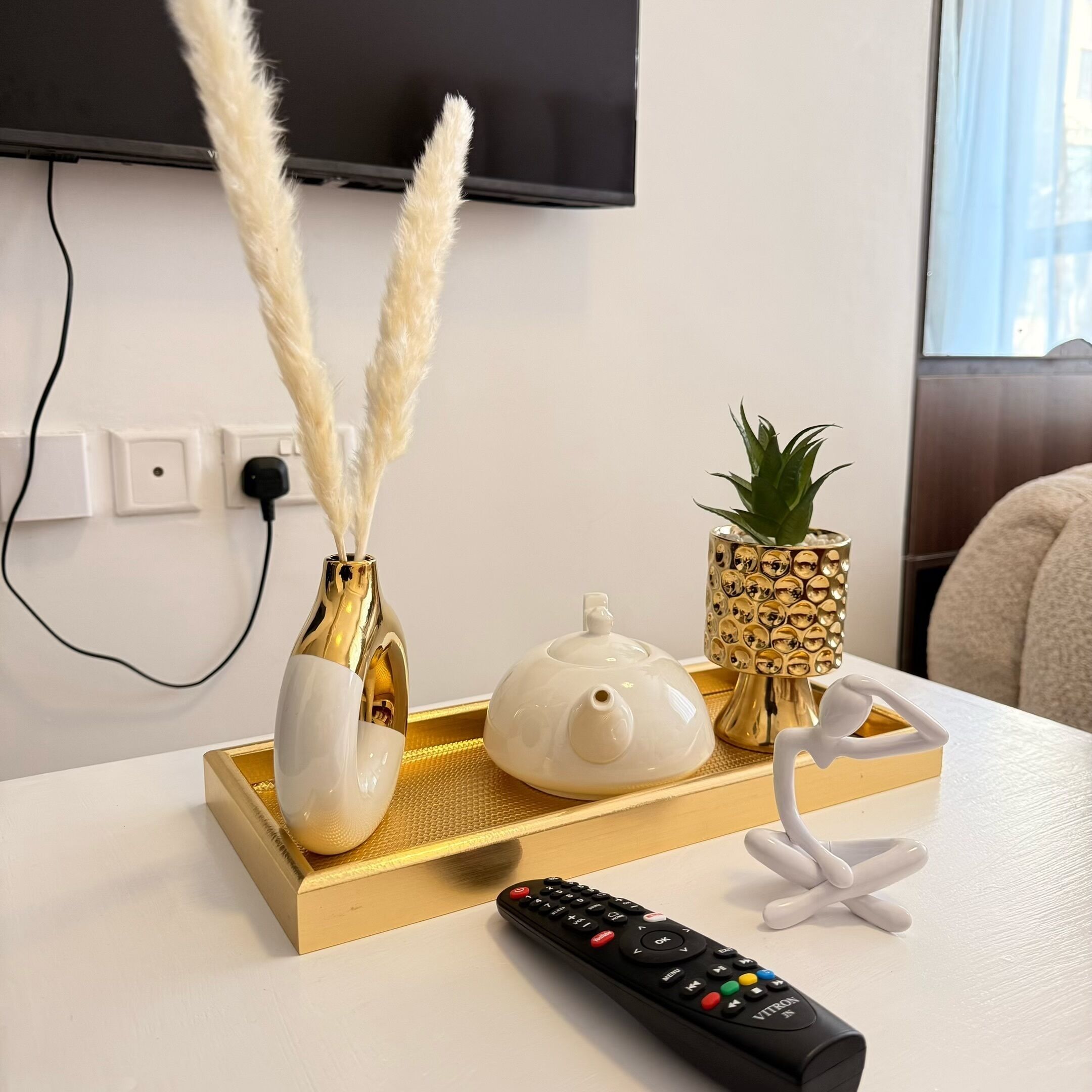 A white table with a gold tray holding a white teapot, two white vases with golden stems, a pineapple, and a black remote control.