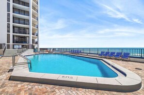 A swimming pool with a clear blue sky, a building with multiple balconies, and a row of blue chairs along the poolside. - Sea Forever at Aquavista 406 E- W PCB - Beachfront (Panama City Beach)