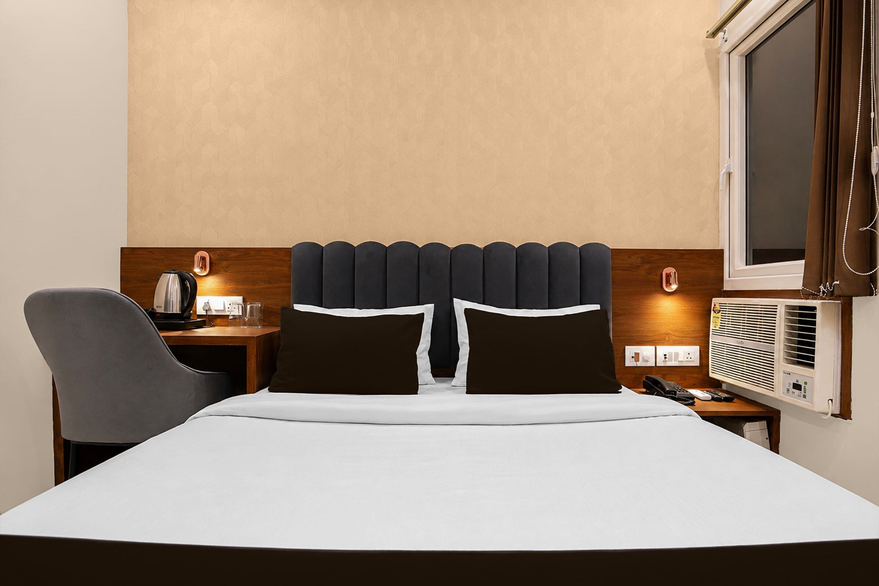 A hotel room with a bed, a desk with a kettle, a chair, a bedside table with a phone, and a window with curtains.