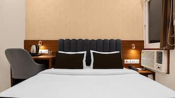 A hotel room with a bed, a desk with a kettle, a chair, a bedside table with a phone, and a window with curtains.