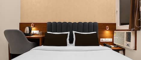 A hotel room with a bed, a desk with a kettle, a chair, a bedside table with a phone, and a window with curtains.