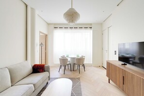 A modern living room with a sofa, a dining table with chairs, a television on a wooden cabinet, and a large window with curtains. - Modern a/c - 1bdr/4p Flat - Montorgueil (Paris)