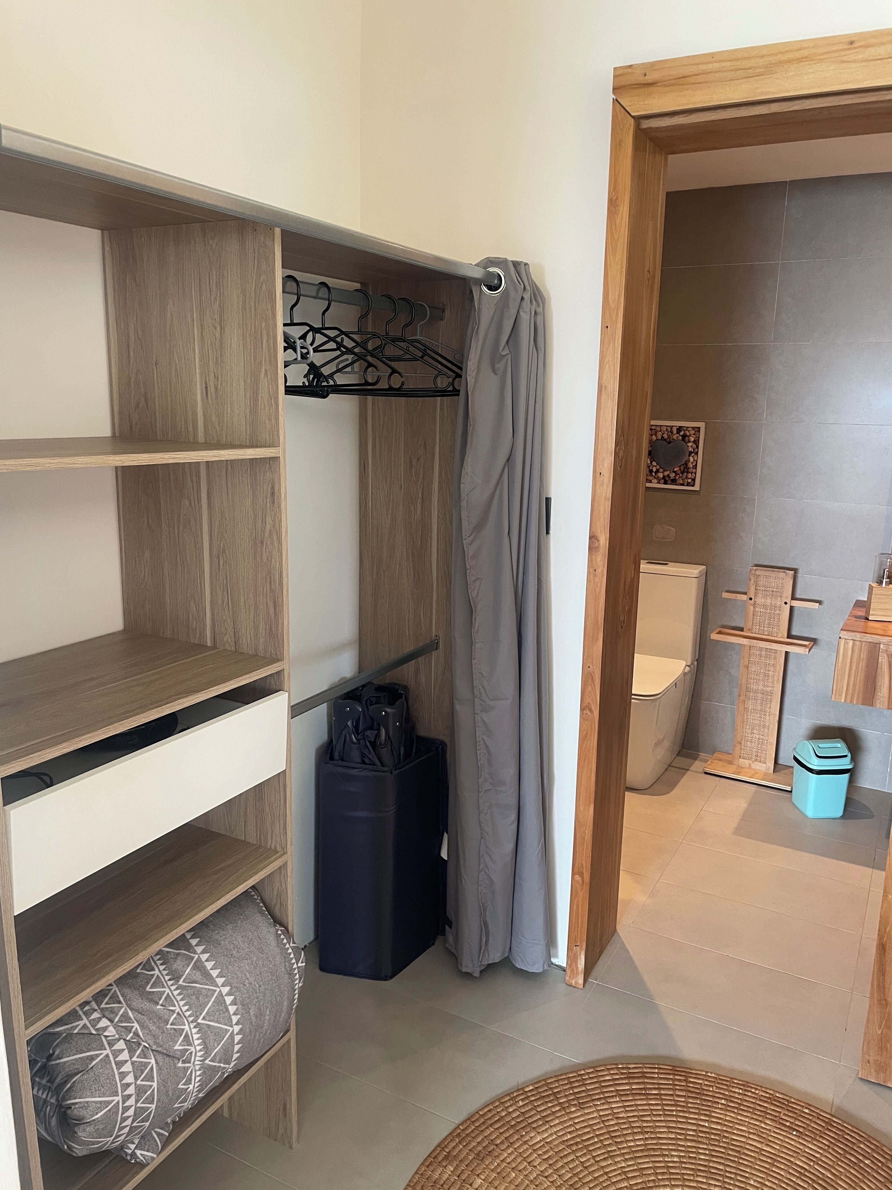 A compact room with a wooden wardrobe, a hanging coat rack, a grey curtain, a small trash bin, and a wicker basket.