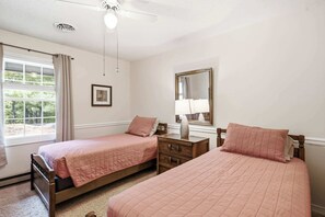 Ein Schlafzimmer mit zwei Betten, einem Fenster mit Vorhängen, einem Deckenventilator, einem Nachttisch und einem Spiegel.