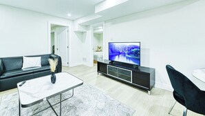 Un salon moderne avec un canapé noir, une table basse en marbre, un téléviseur posé sur un meuble noir et un coin repas avec une table blanche et des chaises noires.