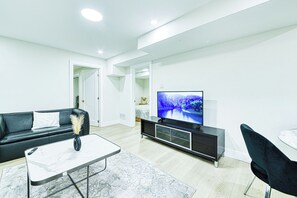 Ein modernes Wohnzimmer mit einer schwarzen Couch, einem Marmortisch, einem Fernseher auf einem schwarzen Lowboard und einem Essbereich mit einem weißen Tisch und schwarzen Stühlen.