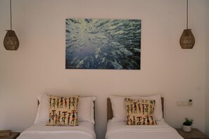 Ein Zimmer mit zwei Betten, jedes mit gemusterten Kissen, einem an der Wand hängenden abstrakten Gemälde und zwei hängenden Korbstühlen.