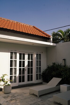 Un bâtiment avec un toit recouvert de tuiles, une façade blanche et une grande porte-fenêtre coulissante. On aperçoit, au premier plan, un pot de fleurs et un fauteuil de jardin en osier.