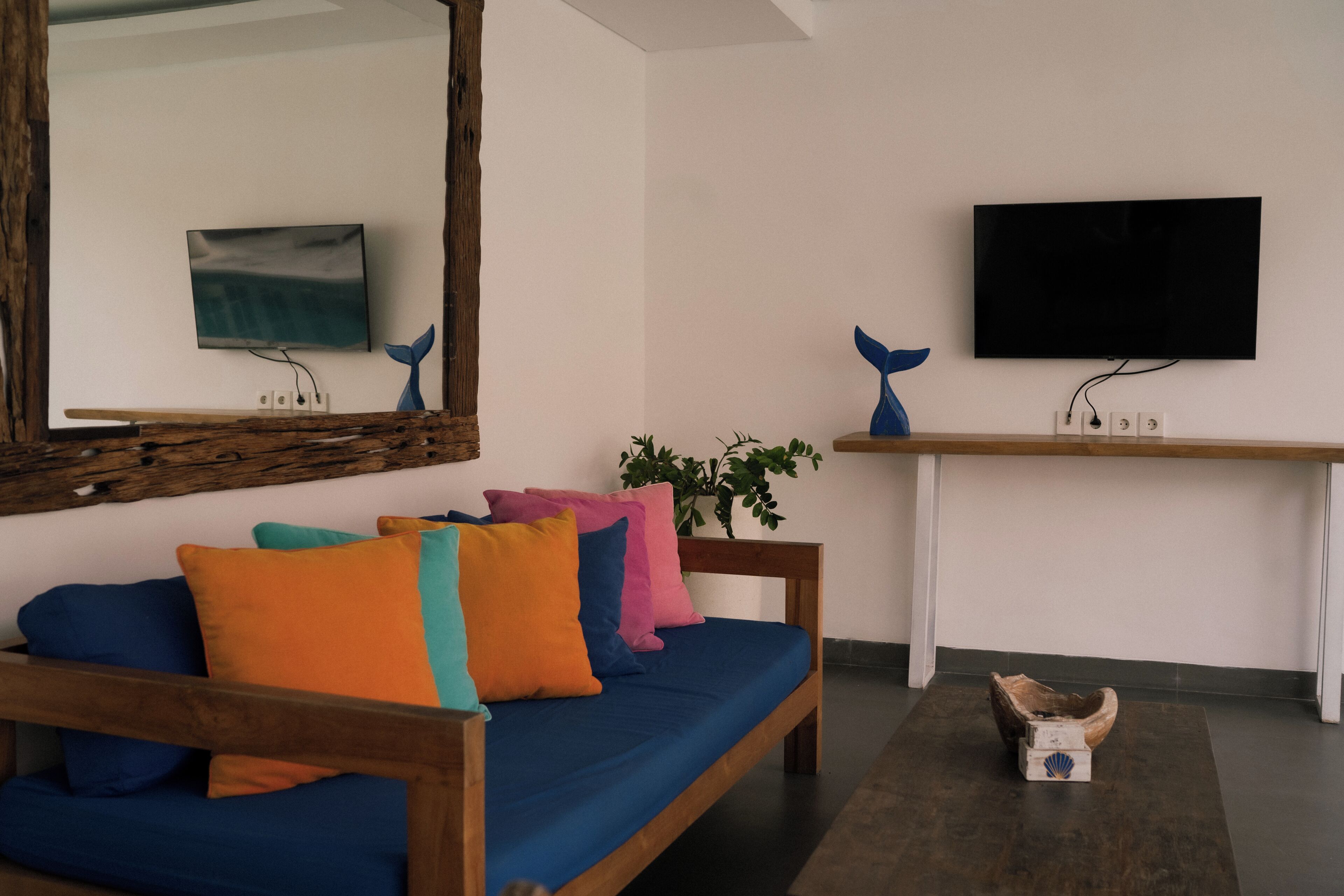 A modern living room with a wooden sofa, colorful cushions, a large mirror, a flat-screen TV, and a wooden side table with a plant.