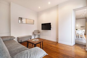 A modern living room with a sofa, a small table, a chair, a flat-screen TV mounted on the wall, and wooden flooring. - Terrace Apartment in Heart of St-Germain-des-Prés (Paris)