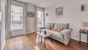 Ein Wohnzimmer mit einer Couch, einem Tisch und einer Lampe. Es gibt Fenster mit Vorhängen, eine Heizung und Kunstwerke an den Wänden.