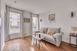 Ein Wohnzimmer mit einer Couch, einem Tisch und einer Lampe. Es gibt Fenster mit Vorhängen, eine Heizung und Kunstwerke an den Wänden.