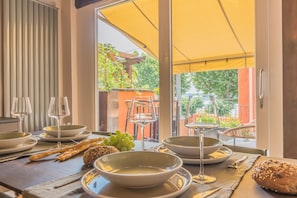 A dining table set with plates, glasses, bread, and grapes, with a view of greenery and a yellow awning. - La Corte dei Laghi. Luxury homestay in the heart of the Italian Lake District. (Pusiano)