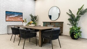 A dining area with a brick wall, a large painting, a round mirror, and potted plants.