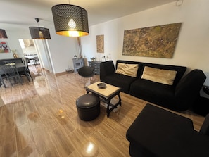 A modern living room with a black sofa, a coffee table, and a black ottoman. There is a dining area with a table and chairs, and a kitchen in the background. - CASA DO PINHEIRO, near Olhos de Água River Beach and the Lake Azul (Pé de Cão)