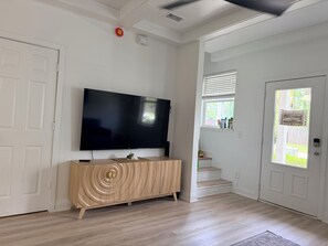 Una habitación con una televisión de pantalla plana montada en la pared, una mesita de madera, una escalera y una puerta con ventanal.