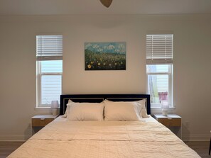 A bedroom with a bed, two bedside tables, a painting above the bed, and two windows with blinds. - Happy Turtle NEW Renovation• Jacuzzi•Grill (Panama City Beach)