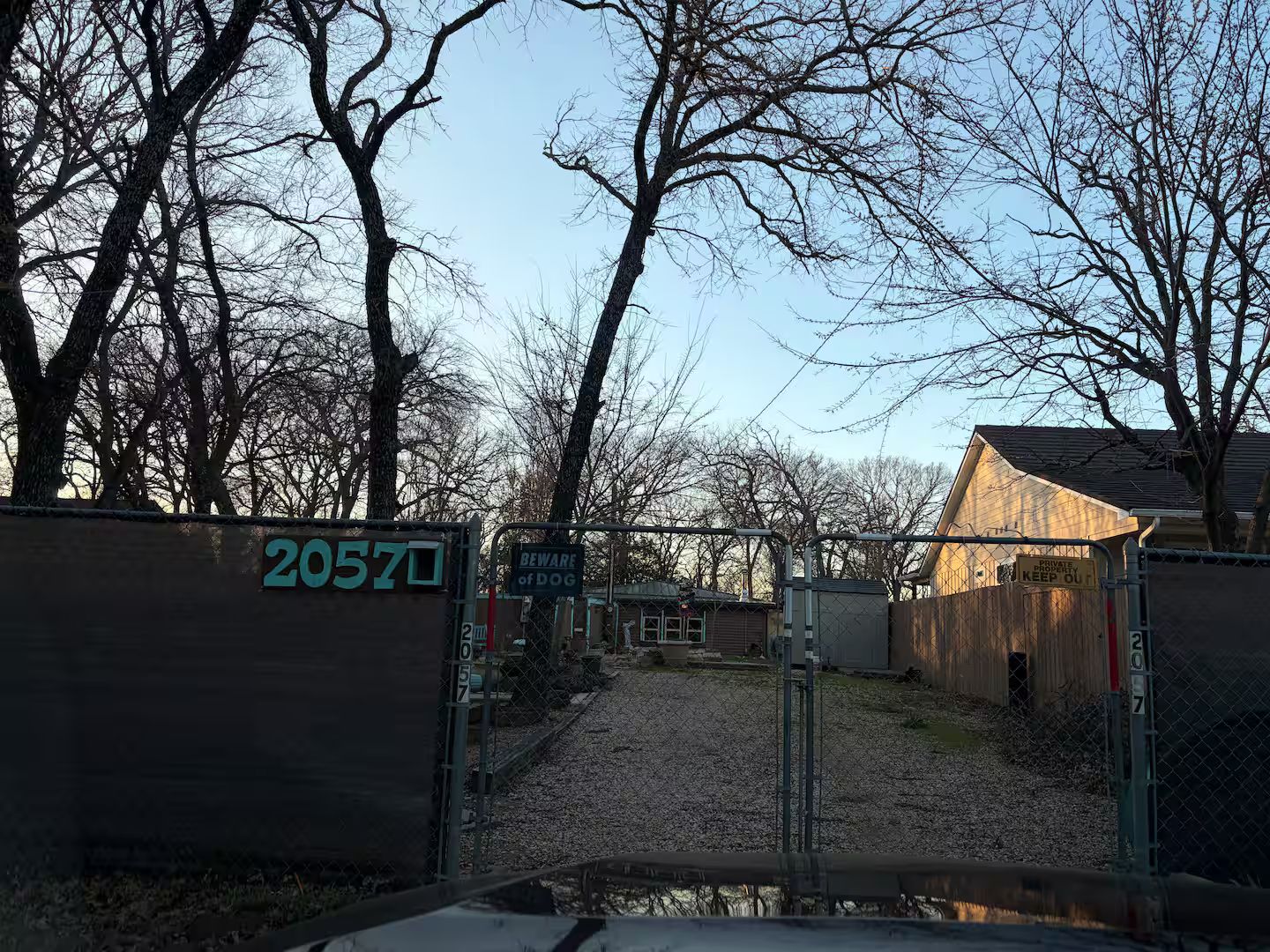 Une entrée de propriété fermée par un portail, avec une plaque indiquant le numéro « 20570 » et un panneau d’avertissement « Attention, chien ».