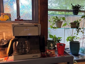 Una cafetera sobre una mesa con vistas a la ventana, una jarra de café, un paquete de Starbucks y plantas en macetas.