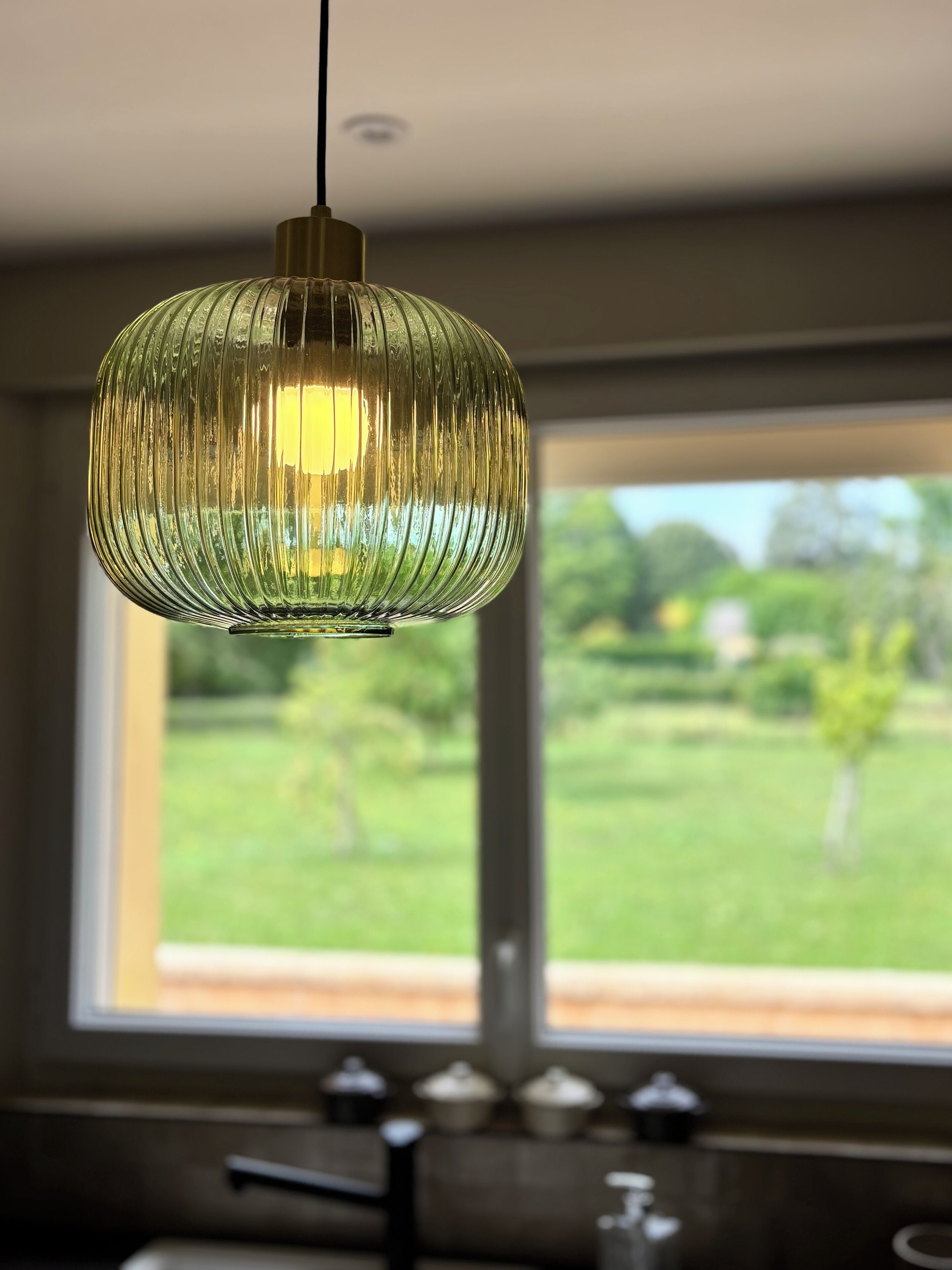 Un luminaire suspendu au design strié, une fenêtre donnant sur un paysage verdoyant et un comptoir de cuisine avec des placards.