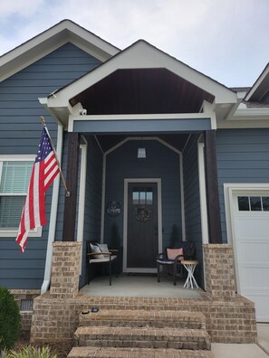 Ein Haus mit dunkelblauer Fassade, einer Veranda mit zwei Stühlen und einem kleinen Tisch, sowie einer amerikanischen Flagge.