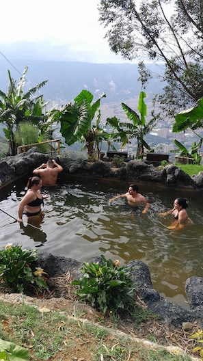 A natural hot spring with people relaxing in the water, surrounded by lush greenery and a view of distant buildings.