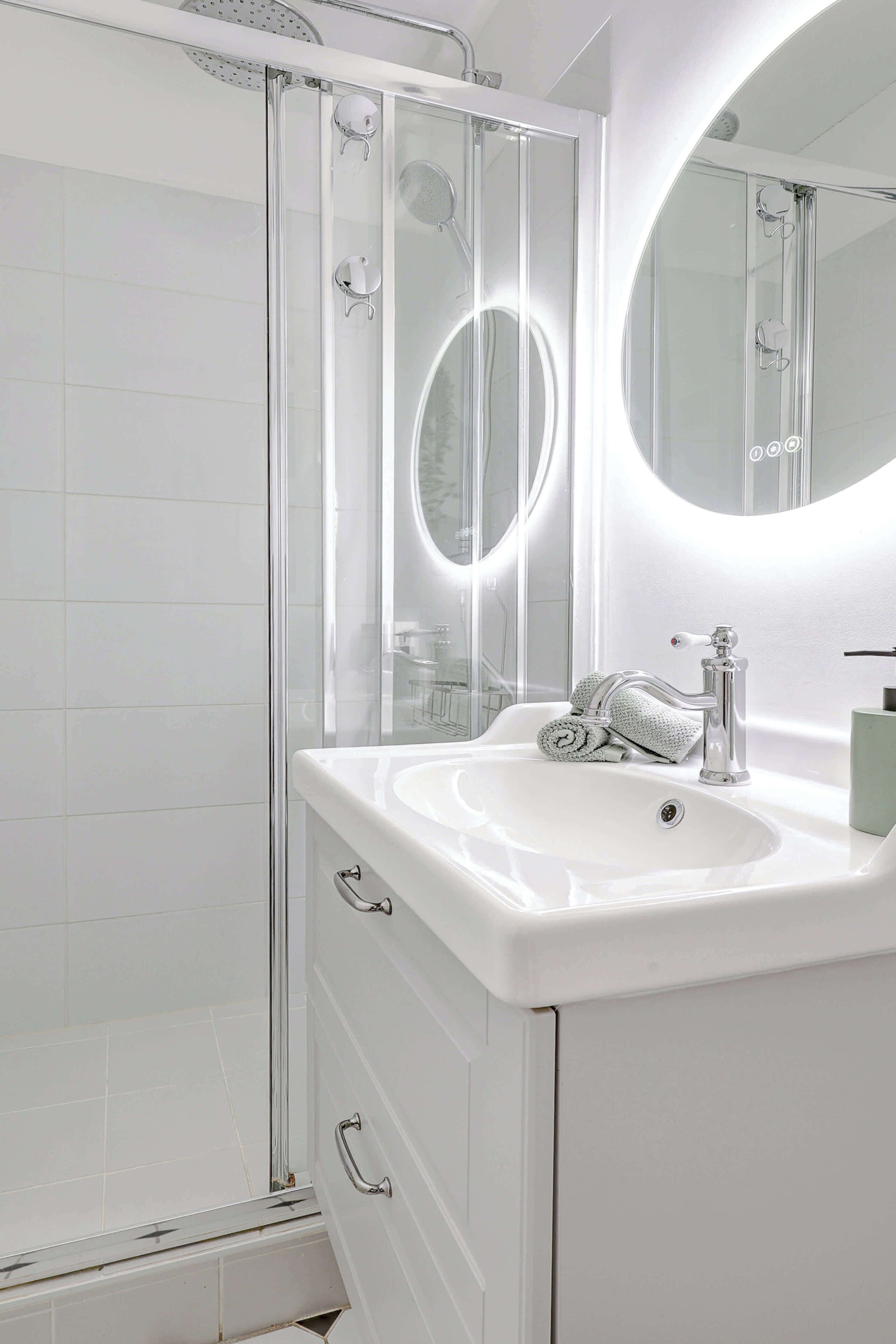 Ein modernes Badezimmer mit einer einteiligen Duschkabine, einem weißen Waschbecken und einem großen Spiegel mit runder Beleuchtung.