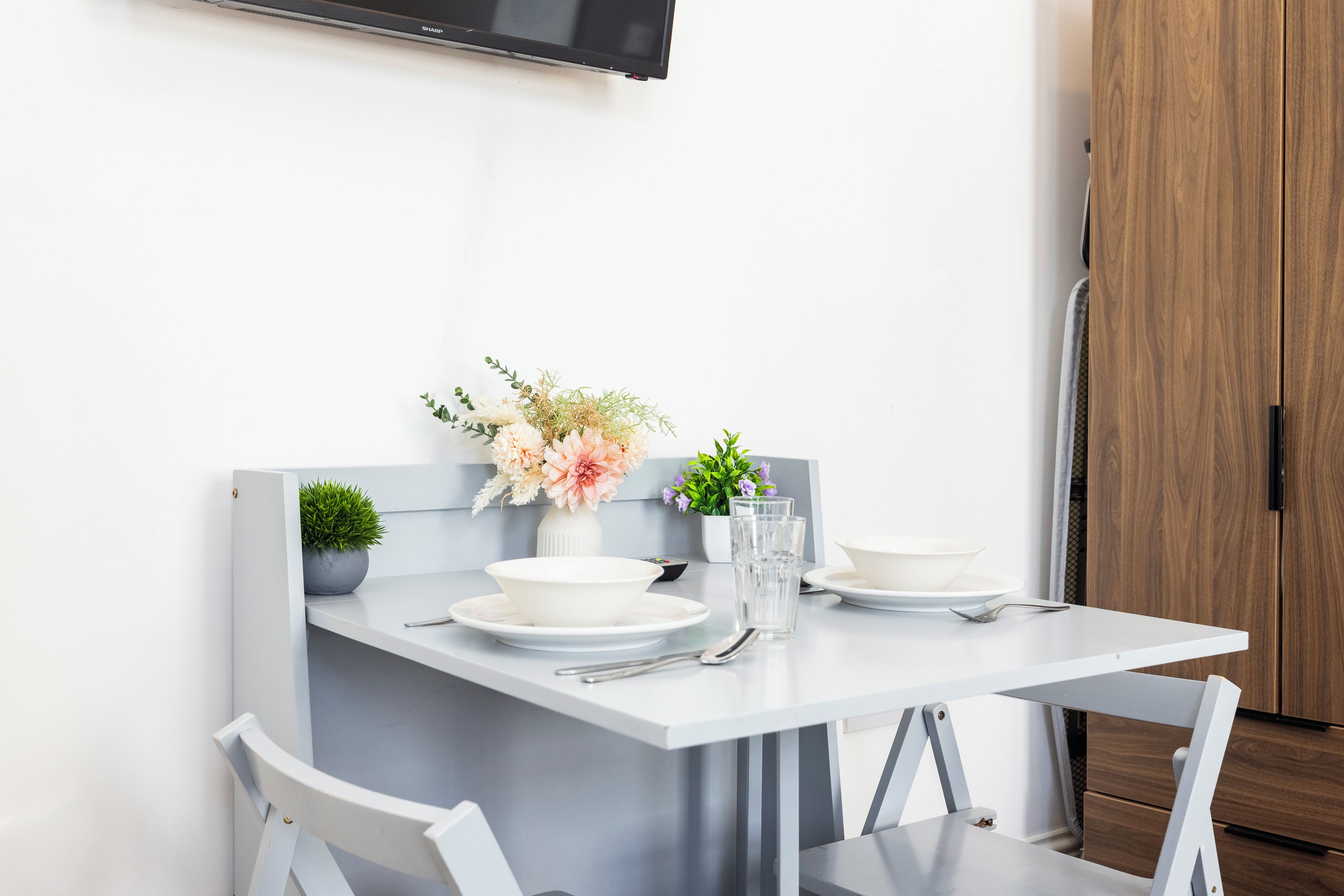 A modern dining area with a white table set for two, a small potted plant, and a vase with flowers.