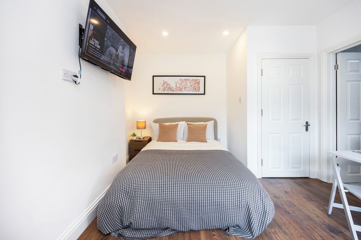 A bedroom with a bed, a TV mounted on the wall, a bedside table with a lamp, and a framed picture on the wall.