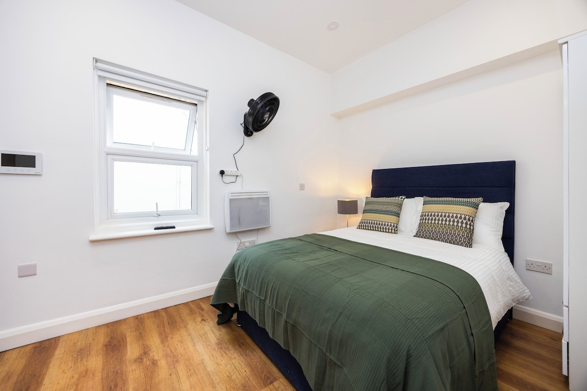 A bedroom with a bed, a window, a wall-mounted heater, and a ceiling fan.